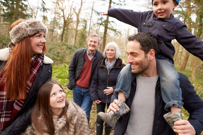 A joyful family strolls through the woods, warmly dressed and surrounded by towering trees. A man carries a child on his shoulders, while a woman and a girl smile at the vibrant canopy above. Two older adults stand behind, exemplifying happiness and security reminiscent of an attentive health insurance service in NJ.