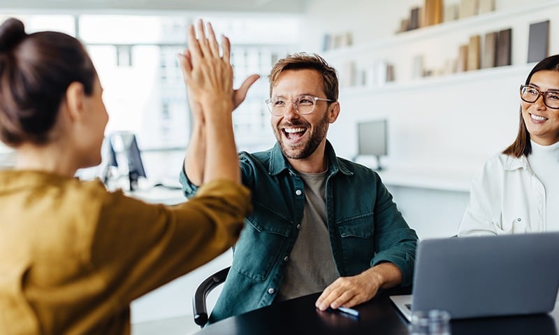 A man with glasses smiles, giving a high-five to someone in a mustard-colored top, while another person in a white jacket sits nearby, smiling. In their modern office setting, home to a leading health insurance broker in New Jersey, shelves and a computer can be seen in the background.