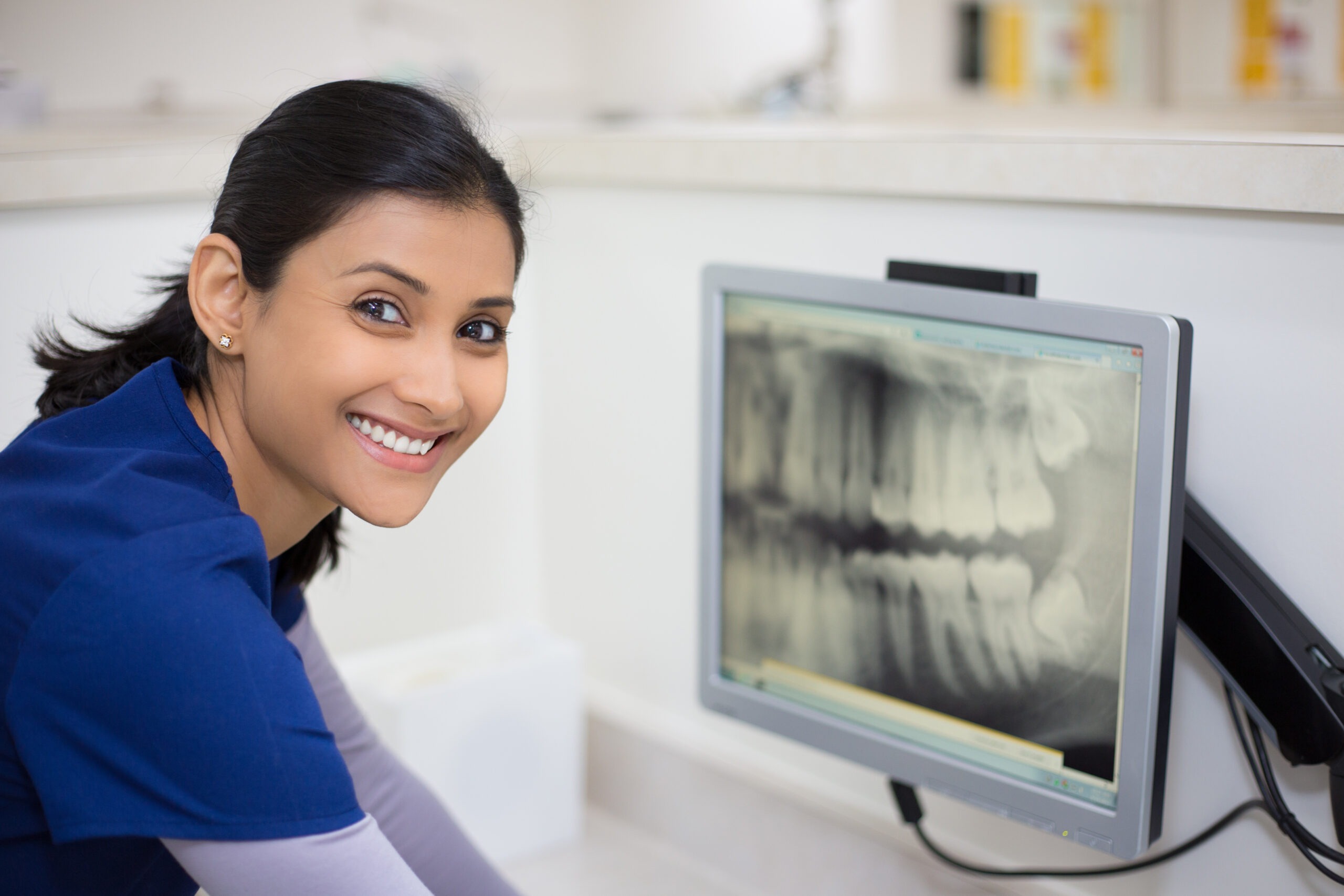 A smiling person in a blue uniform is seated next to a computer monitor displaying a dental X-ray, embodying the professional care you're assured of when consulting with a health insurance specialist in NJ. The background reveals an office or clinic setting, reflecting a commitment to patient well-being.