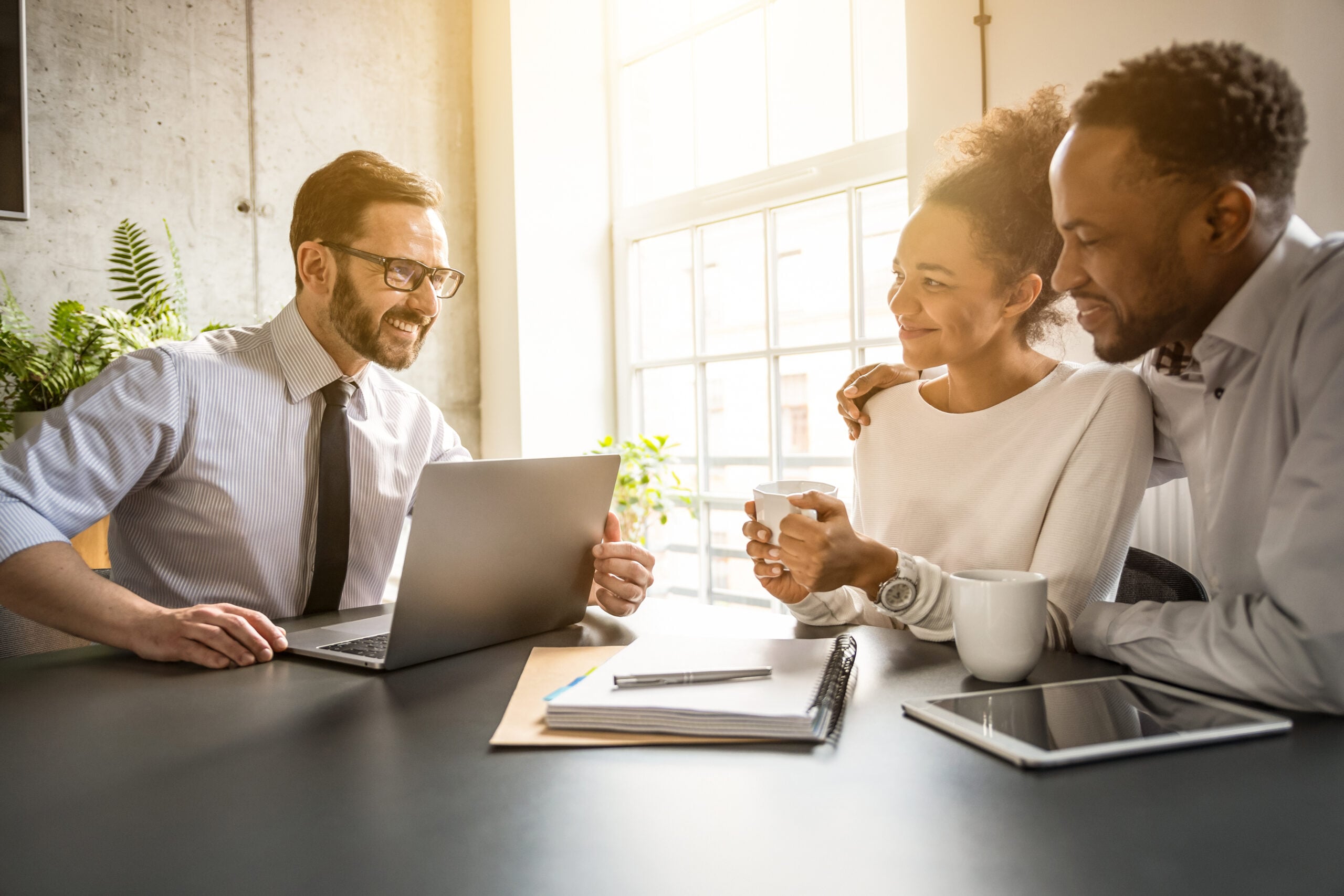 A health insurance agent in NJ sits at a desk with a laptop, smiling and talking to a couple who are holding coffee cups. Papers, a pen, and a tablet accompany the scene as sunlight streams through the window.