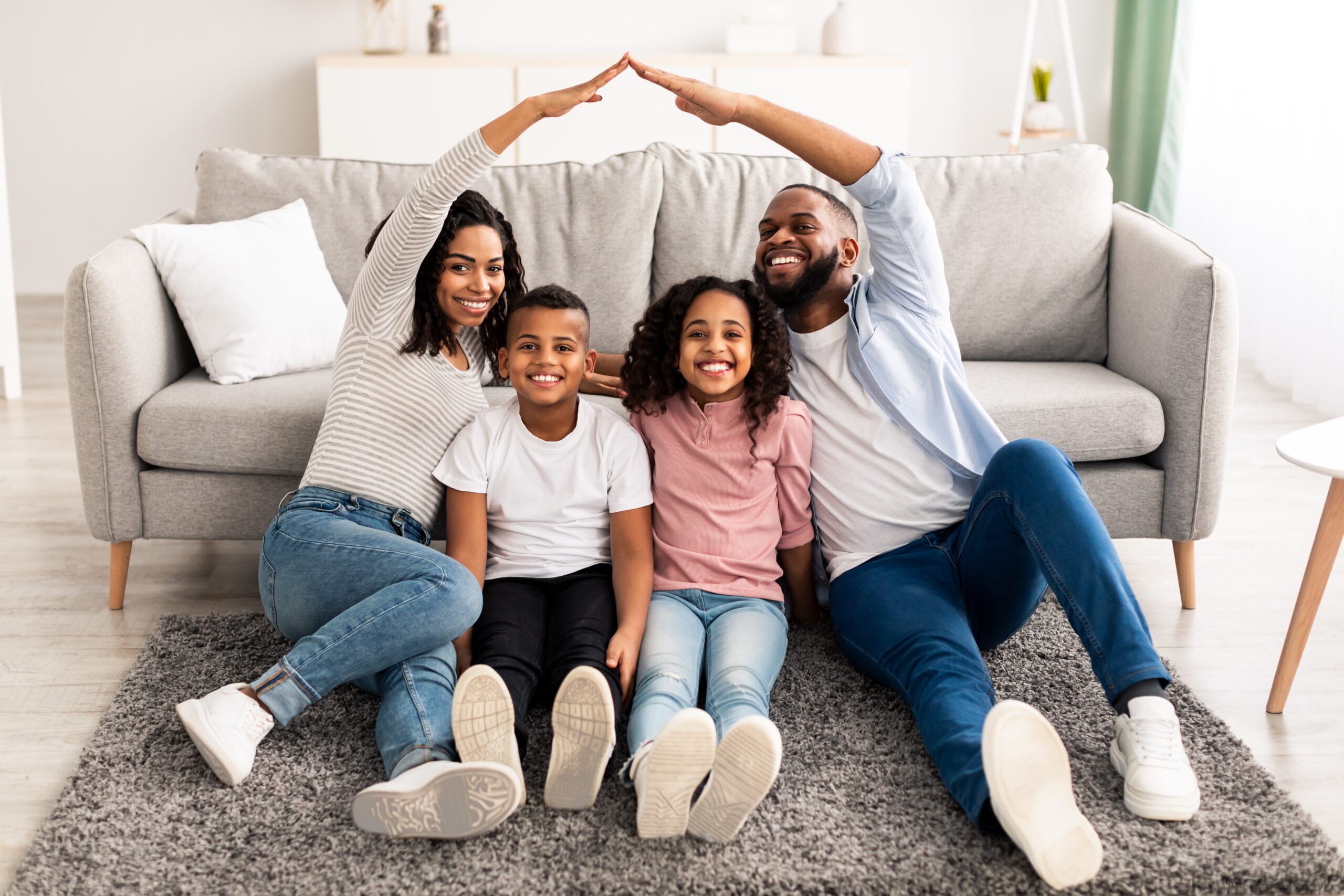 A happy family of four sits on a gray rug in front of a couch, enjoying peace of mind thanks to their health insurance service in NJ. The parents form a roof shape with their arms above their children’s heads. Everyone is smiling, creating a warm and joyful atmosphere in the well-lit living room.