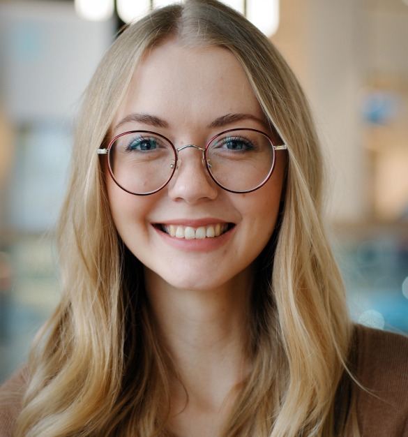 A young woman with long blonde hair and glasses smiles at the camera, radiating a professional charm. In her brown top, she embodies the dedication of a health insurance specialist in NJ. The softly blurred indoor background adds to her approachable aura.