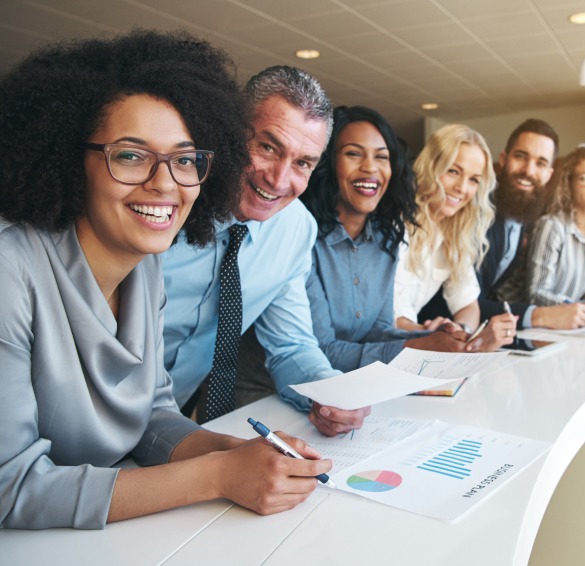 A group of six diverse professionals sits at a table smiling while holding documents with charts. They appear engaged and friendly, discussing their roles as a health insurance broker in New Jersey in a modern office setting.