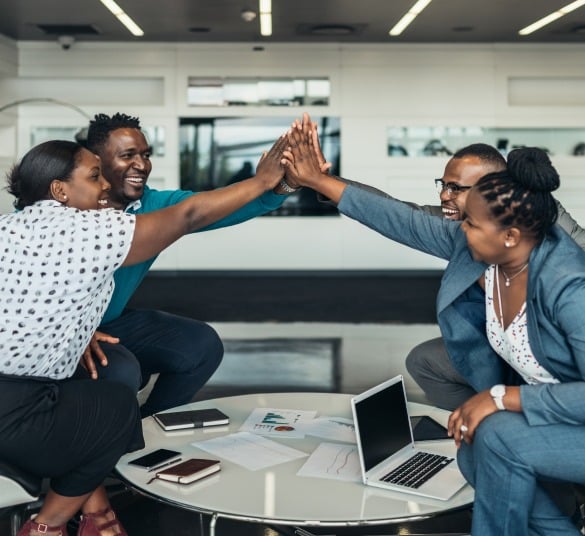 A group of four professionals, including a health insurance specialist in NJ, sit around a table in an office, smiling and high-fiving each other. A laptop, papers, and smartphones are on the table, indicating a collaborative and successful meeting.