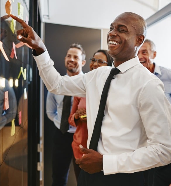 A group of business professionals, including a health insurance broker in New Jersey, are smiling and engaged in a discussion. One person is pointing at sticky notes on a board, leading the conversation. They appear to be in a modern office setting.
