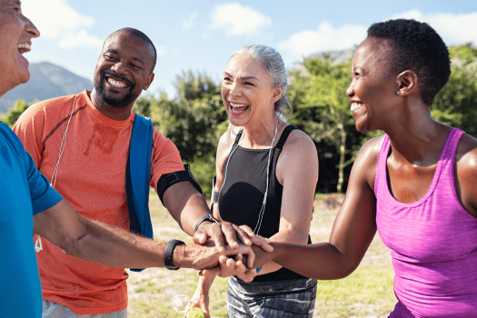 A diverse group of four adults, seemingly led by a health insurance specialist in NJ, laugh and put their hands together in a circle outdoors. Dressed in sports attire, they enjoy exercising on a sunny day with trees in the background.
