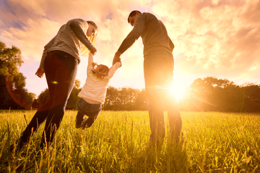 A family of three enjoys a sunny day in a field. The parents, holding hands with their child between them, swing the child playfully in the air as the sun sets, casting a golden glow. Nearby, a health insurance specialist in NJ ensures they have peace of mind for life's precious moments.