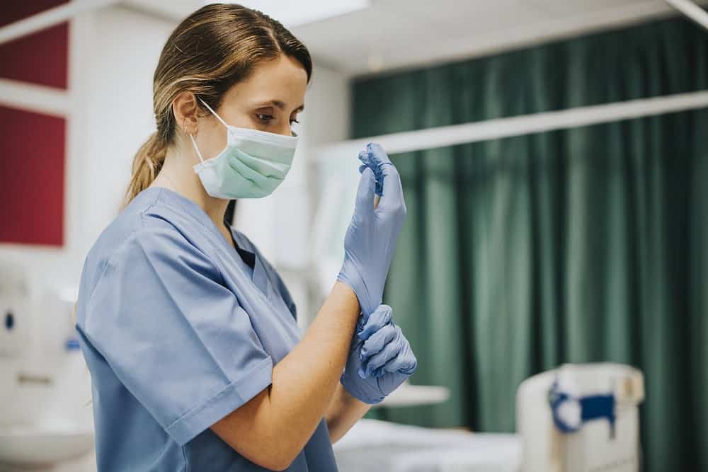 A healthcare worker, resembling a diligent health insurance specialist in NJ, dons blue gloves in a medical setting. Scrubs and a face mask complete the look, while a green curtain and medical equipment form the backdrop of this crucial healthcare environment.