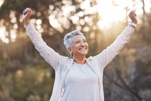 A joyful person with short gray hair raises their arms triumphantly outdoors, exuding the positivity of finding a great health insurance service in New Jersey. They are smiling, wearing a light-colored jacket and shirt, with sunlight filtering through trees in the background, creating a warm, serene atmosphere.