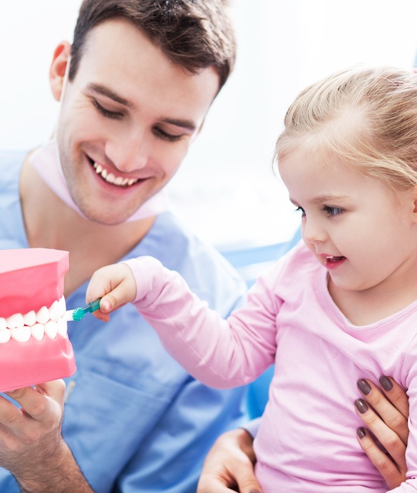 A dentist shows a young girl how to brush teeth using a large model of a mouth. The girl, wearing a pink shirt, is holding a toothbrush and smiling as she learns. With the support of health insurance services in NJ, both can enjoy these essential health practices with peace of mind.