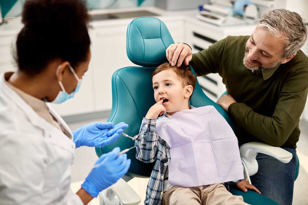 A young boy sits in a dentist chair with a dental bib, touching his mouth, while a dentist in a white coat and mask holds dental tools. An older man, possibly the family's health insurance specialist in NJ, sits beside the boy, gently comforting him. They are in a dental office.