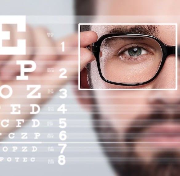 A close-up of a man, possibly a health insurance broker in New Jersey, adjusting his black-rimmed glasses while focusing on an eye chart. The chart is slightly blurred in the background, displaying rows of letters in descending sizes.