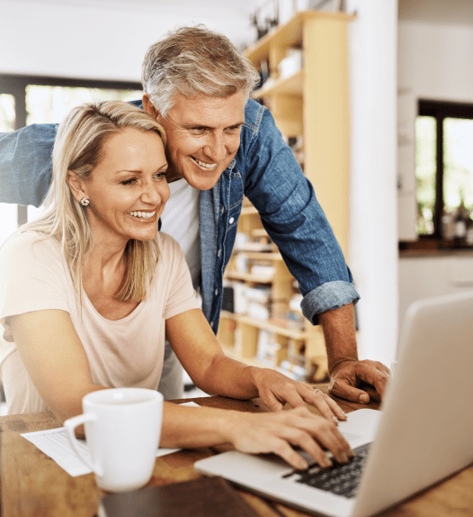 A smiling couple in a cozy room looks at their laptop screen together, exploring options with a health insurance broker in New Jersey. The woman is seated, typing, as the man leans over her shoulder. A coffee mug sits nearby on a wooden table with shelves of books and decor in the background.