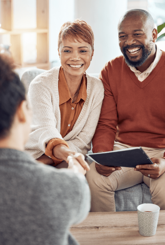 A smiling couple sits on a couch in a warmly lit room, engaging in conversation with a health insurance specialist in NJ. The woman reaches forward to shake hands with the third person, partially visible. The man holds a black folder on his lap. A coffee cup is on the table.