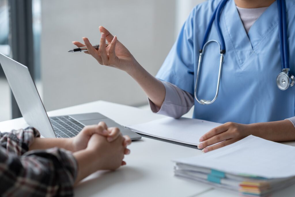 A healthcare professional in scrubs sits at a desk, gesturing with a pen during a discussion with someone whose hands are clasped together. A laptop and a stack of papers rest on the desk as they consult about options with their health insurance specialist in NJ.