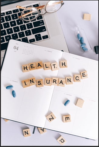 A flat lay image features a laptop, open planner, and eyeglasses. Wooden letter tiles spell "HEALTH INSURANCE" on the planner. Blue pills are scattered nearby, evoking healthcare planning themes reminiscent of consulting a health insurance broker in New Jersey.