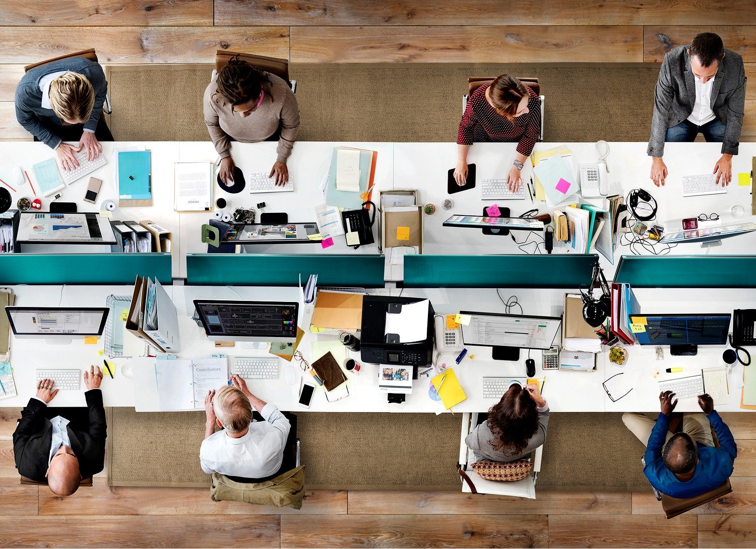 Overhead view of office workers sitting at desks, working on computers and using phones. Desks are organized in two rows with papers, notebooks, and office supplies scattered around.