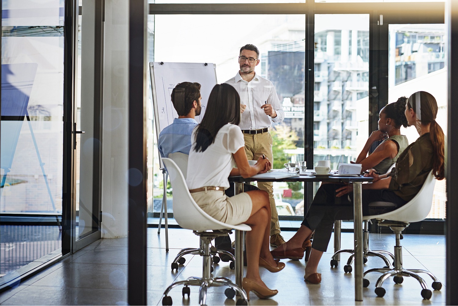 A man stands by a flip chart giving a presentation to four colleagues seated around a glass table in a modern office with large windows.