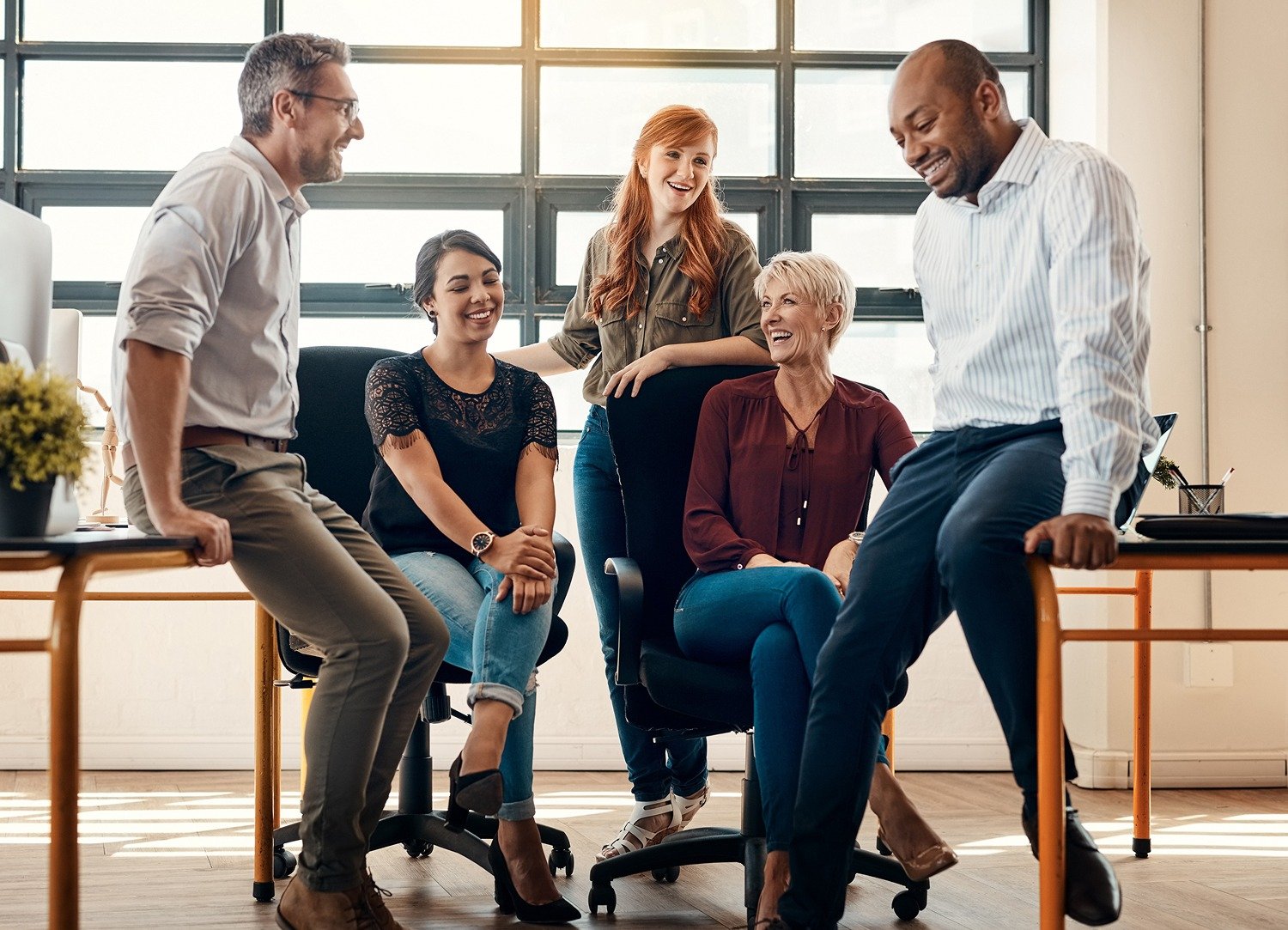 Five colleagues, three women and two men, casually chat and smile together in a bright office space with large windows, creating a relaxed and friendly atmosphere.